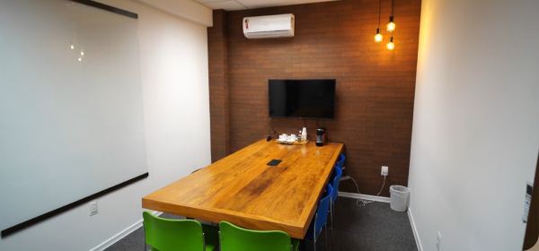 Sala de reunião do Foccus Workplace com mesa de madeira maciça, televisor na parede e iluminação pendente.
